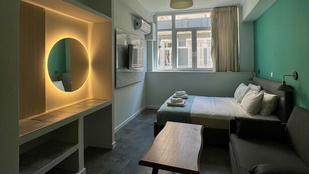 A compact hotel room with a bed, a sofa, a small table, a round mirror, and a TV. - Monastiraki 101 @SuperHost Hub (Athina)