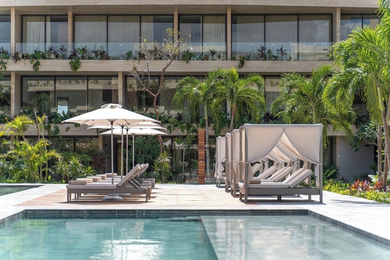 Area piscina con lettini e ombrelloni, immersa nel verde e affiancata da un edificio moderno con ampie finestre.