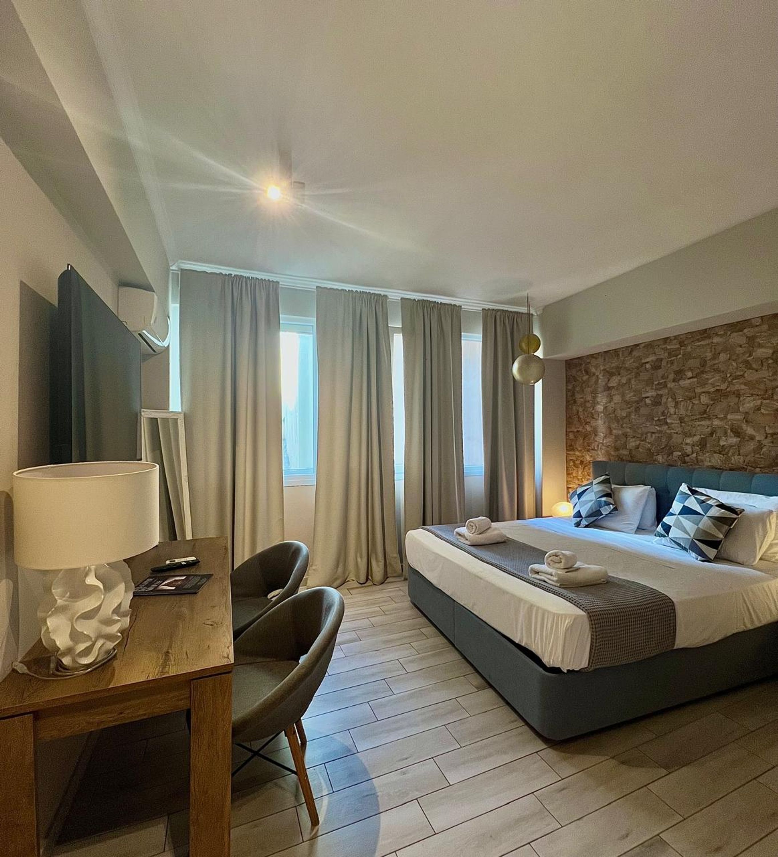 A hotel room with a bed, a desk with a lamp, a chair, and a stone wall.