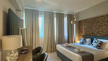 A hotel room with a bed, a desk with a lamp, a chair, and a stone wall.