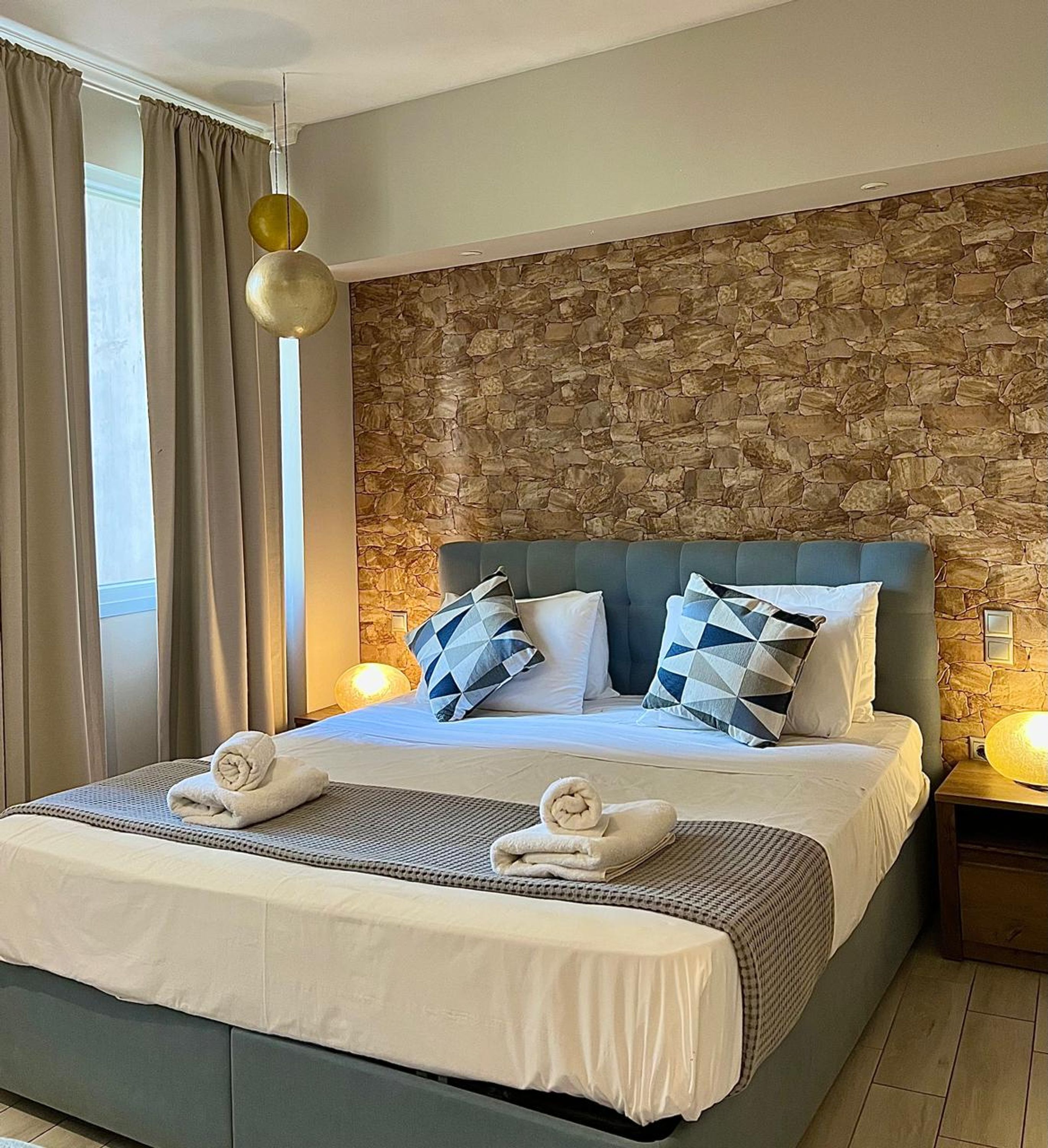 A hotel room with a stone wall, a large bed with a blue headboard, two bedside lamps, and a window with curtains.