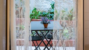 Vista a través de un cortinaje de encaje, mostrando un balcón con mesa y sillas, una planta en maceta y diversas plantas verdes al fondo.