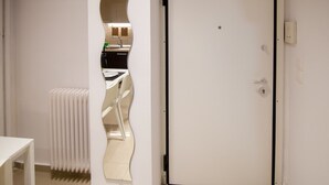 Una stanza con una porta bianca, un termosifone, un tavolo bianco e uno specchio che riflette una zona cucina.