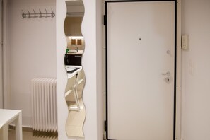 Une pièce avec une porte blanche, un radiateur, une table blanche et un miroir qui reflète un coin cuisine.