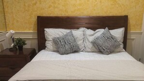 A neatly made bed with a striped bedspread and two patterned pillows, a wooden nightstand with a lamp and a small plant, and a yellow textured wall. - VanDusen Homestay in Vancouver Close to UBC and Public Transits (Vancouver)