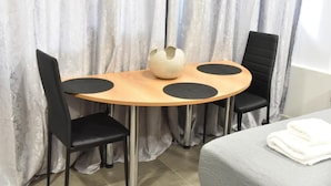 Een kamer met een ronde houten tafel, twee zwarte stoelen, een witte vaas en een bed met een witte deken.