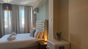 A hotel room with a large bed, a bedside table with a lamp, a small plant, and a window with curtains. - Monastiraki 509 Central Suite (Athina)