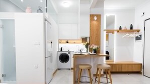 Cuina compacta amb rentadora, zona de dinar petita amb tamborets i prestatge de fusta amb objectes penjats.