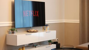 Un salon moderne avec un téléviseur fixé au mur affichant le logo de Netflix, une console blanche agrémentée d’objets décoratifs, une petite maquette de maison et une table basse ronde.