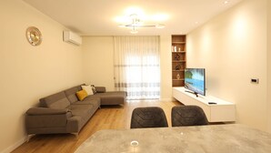 A modern living room with a grey sectional sofa, a dining table with chairs, a TV on a white stand, and a bookshelf. - Grandstay Portside in Durrës (Durrës)