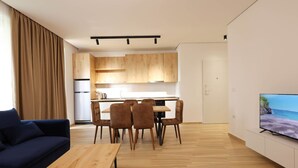 Een moderne woonkamer met een bank, een eettafel en stoelen. In de achtergrond is een keuken met houten kastjes en een koelkast zichtbaar.