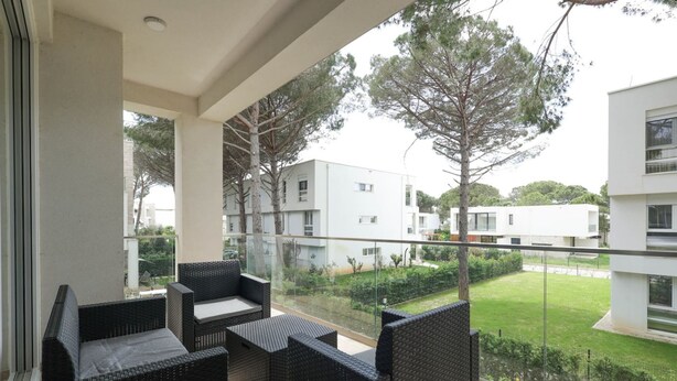 Een balkon met rieten meubilair, een glazen hekwerk en uitzicht op moderne gebouwen en bomen.