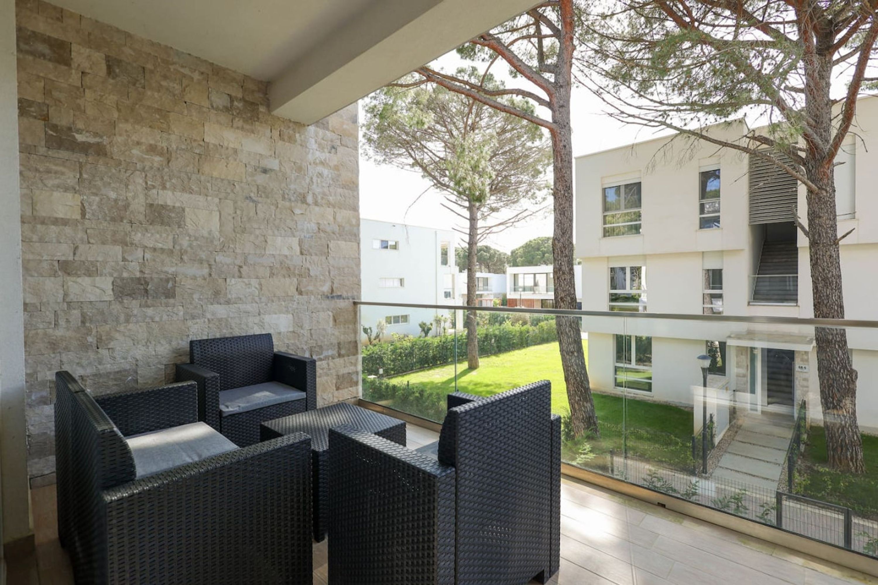 Een balkon met rieten meubilair, een stenen muur en uitzicht op moderne gebouwen en groen.