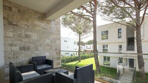 Ein Balkon mit Korbmöbeln, einer Steinmauer und Blick auf moderne Gebäude und Grünflächen.