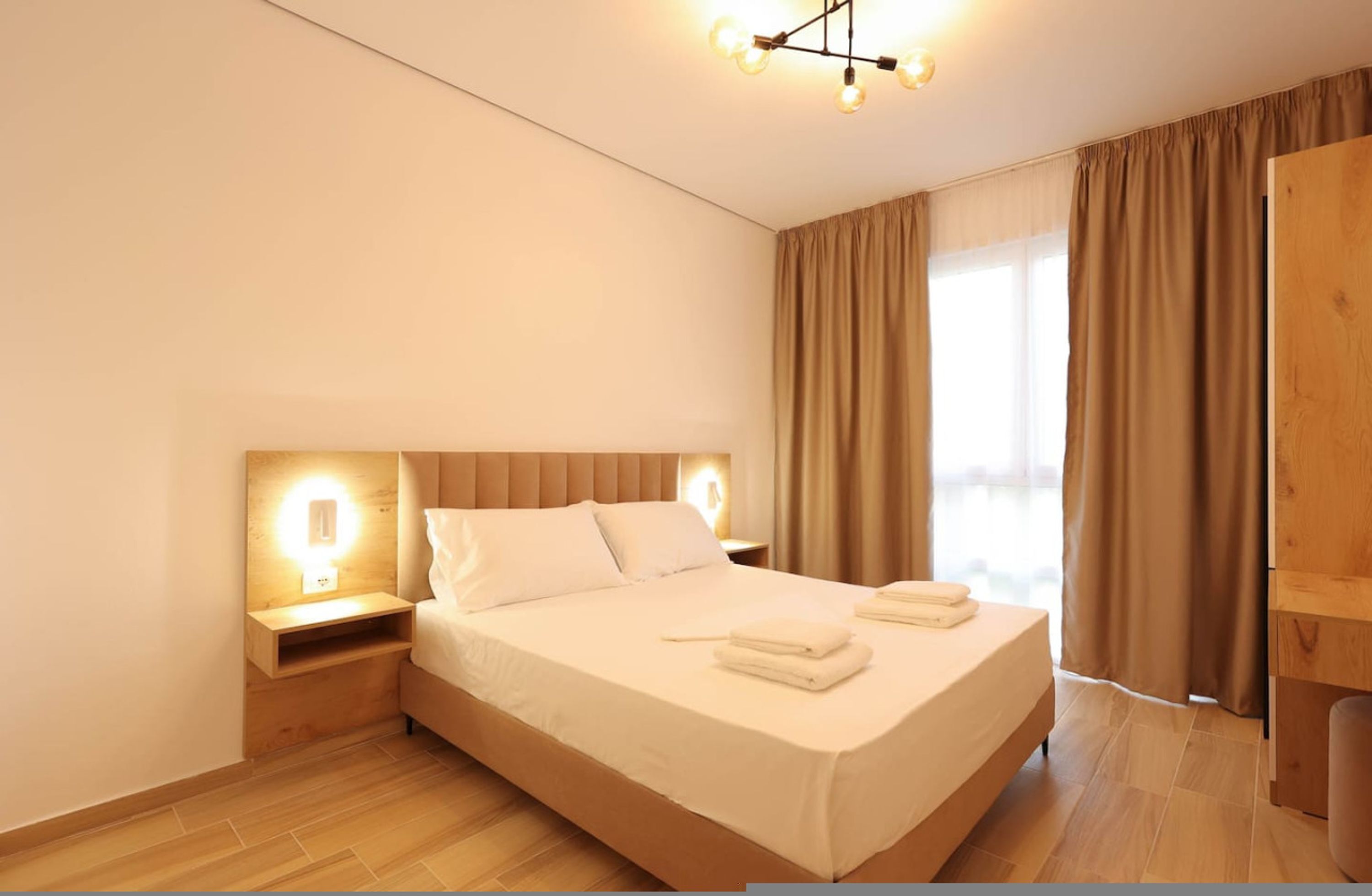 Ett modernt hotellrum med en stor säng, ett träbeklätt sänggavel, sänglampor och utsikt över omgivningarna genom stora fönster.