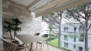 Een balkon met een tafel en stoelen, een plant in pot en uitzicht op een gebouw en bomen.