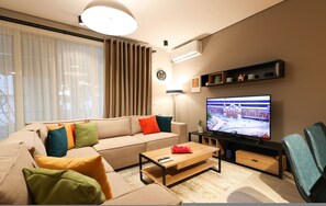 A modern living room with a beige sofa, colorful cushions, a wooden coffee table, a flat-screen TV, and a large window with sheer curtains. - SanPietro Vacation Apartment 011 (Plazhi San Pietro)