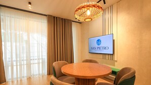 Una habitación de hotel moderna con una mesa redonda de madera, sillas tapizadas en tonos beige y verde, un ventanal con cortinas translúcidas, un televisor empotrado mostrando 'SAN PETRO Vacation Rentals', y una lámpara colgante de fibras naturales.