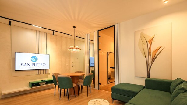 Una sala de estar moderna con un sofá verde, una mesa redonda con sillas y un televisor de pantalla plana mostrando 'SAN PIETRO'.