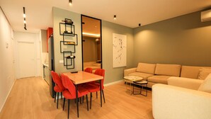 A modern living room with a beige sofa, a dining table with red chairs, and a kitchen area with a white door. - SanPietro Vacation Apartment 026 (Plazhi San Pietro)
