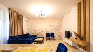 A modern living room with a blue sofa, a small table, and two chairs. There is a TV on a wooden shelf and a ceiling light. - SanPietro Vacation Apartment 032 (Plazhi San Pietro)