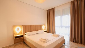 A hotel room with a large bed, two bedside tables with lamps, a window with curtains, and a wooden floor. - SanPietro Vacation Apartment 032 (Plazhi San Pietro)