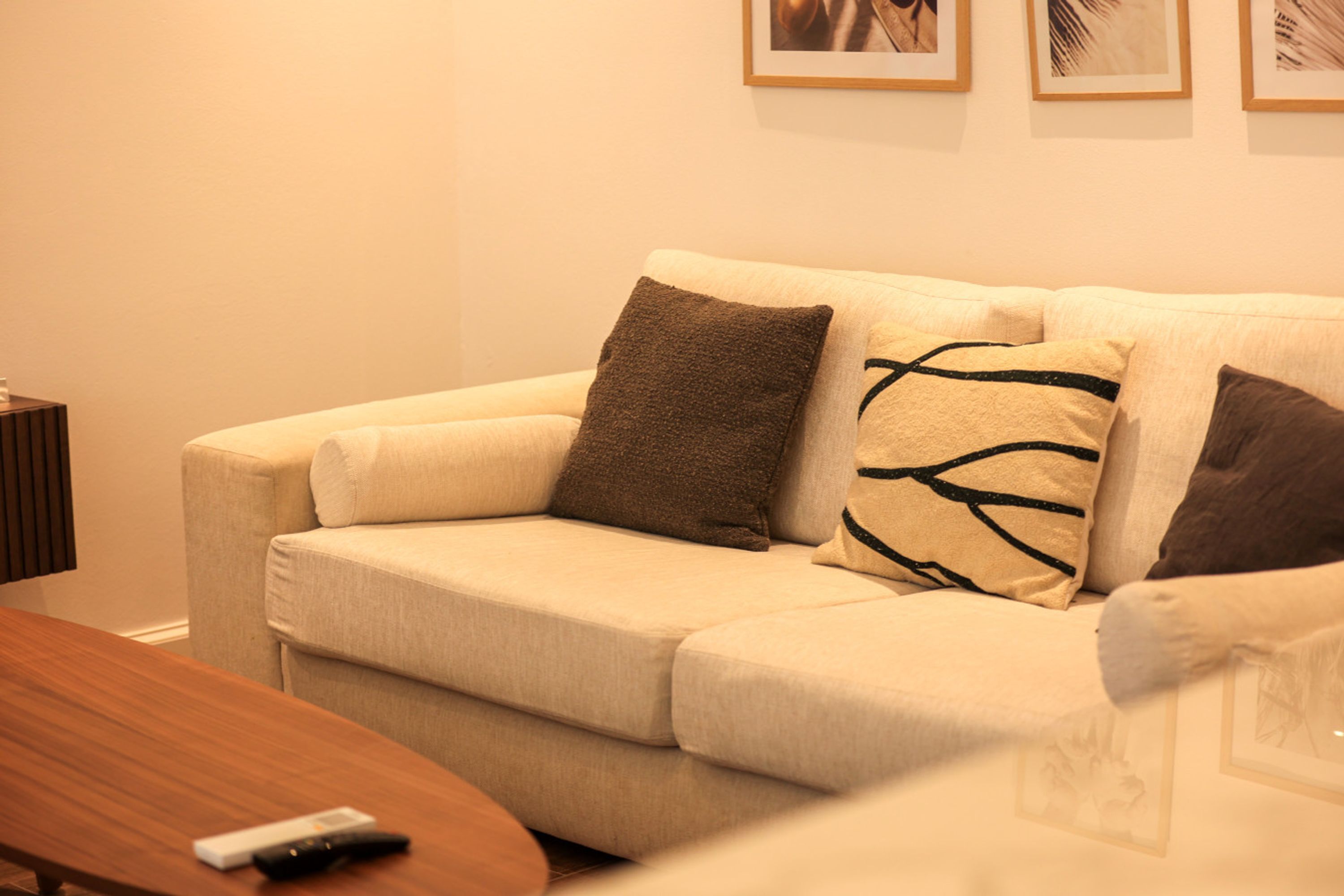 Un sofá beige con dos almohadas, un control remoto sobre una mesa de madera y cuadros enmarcados en la pared.