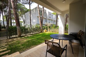 A patio area with a round table and chairs, surrounded by trees and a white building in the background. - SanPietro Vacation Apartment 028 (Plazhi San Pietro)