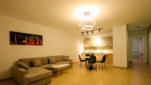 A modern living room with a beige sofa, a small coffee table, and a dining area with a round table and chairs. There is a kitchen visible in the background with wooden cabinets and a white countertop. - SanPietro Vacation Apartment 033 (Durrës)