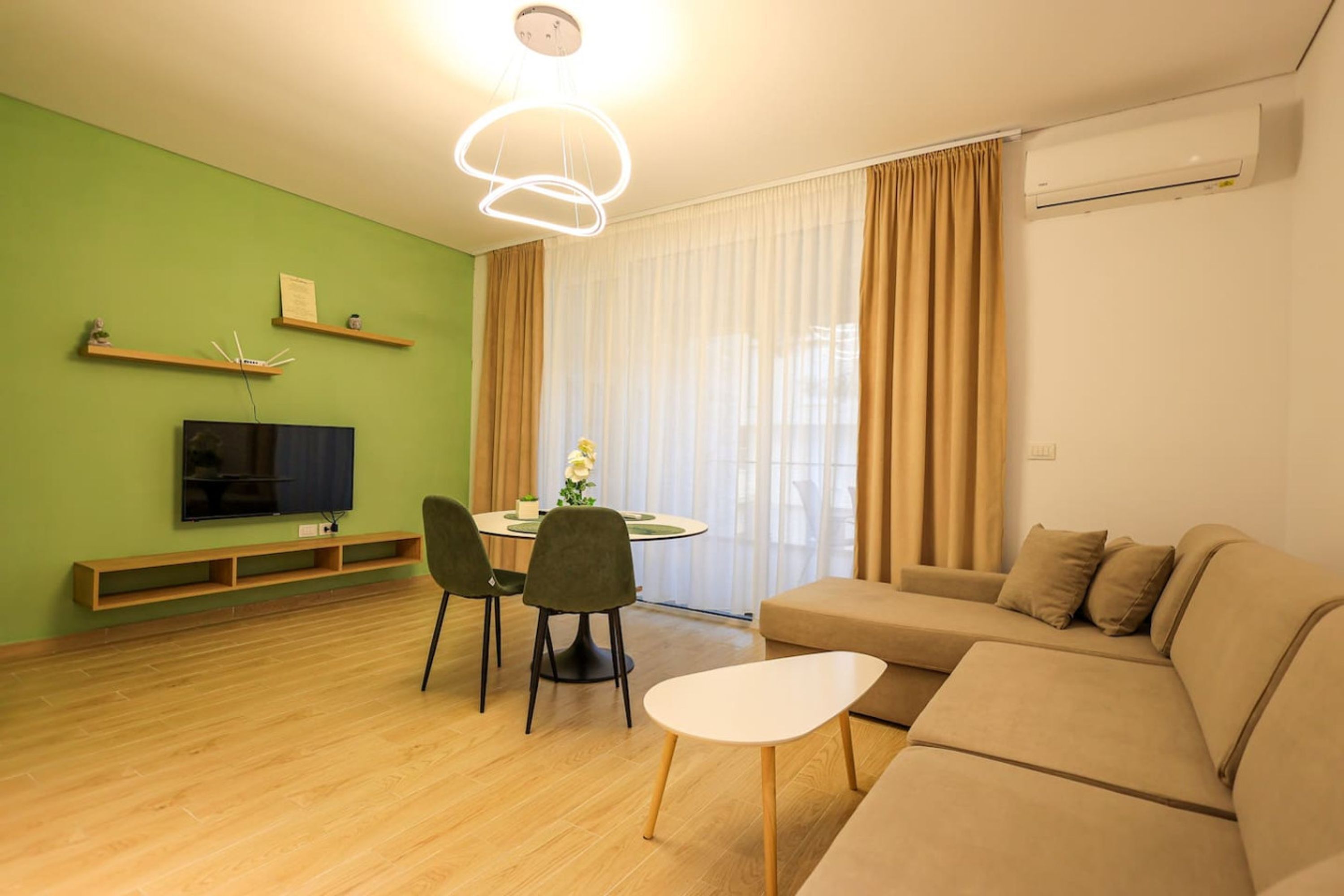 Un salon moderne avec un mur vert, un plancher en bois, un canapé beige, une table à manger avec des chaises, un téléviseur à écran plat et un luminaire suspendu au plafond.