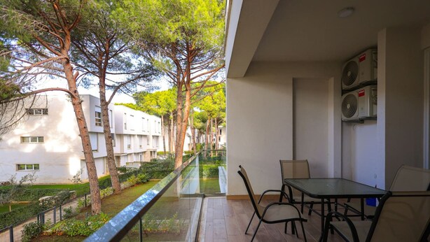 Ein Balkon mit Glastrennwand, der auf einen Garten und ein Gebäude blickt. Dort stehen ein Tisch und Stühle, und eine Wandleuchte ist an der Wand befestigt.