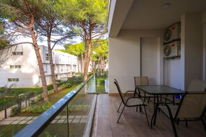 Un balcón con barandilla de vidrio que da a un jardín y un edificio. Hay una mesa con sillas y un ventilador fijado a la pared.