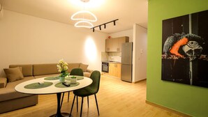 Ein modernes Wohnzimmer mit einem runden Esstisch, grünen Stühlen und einer Couch. Im Hintergrund ist eine Küche mit Kühlschrank und Schränken zu sehen.