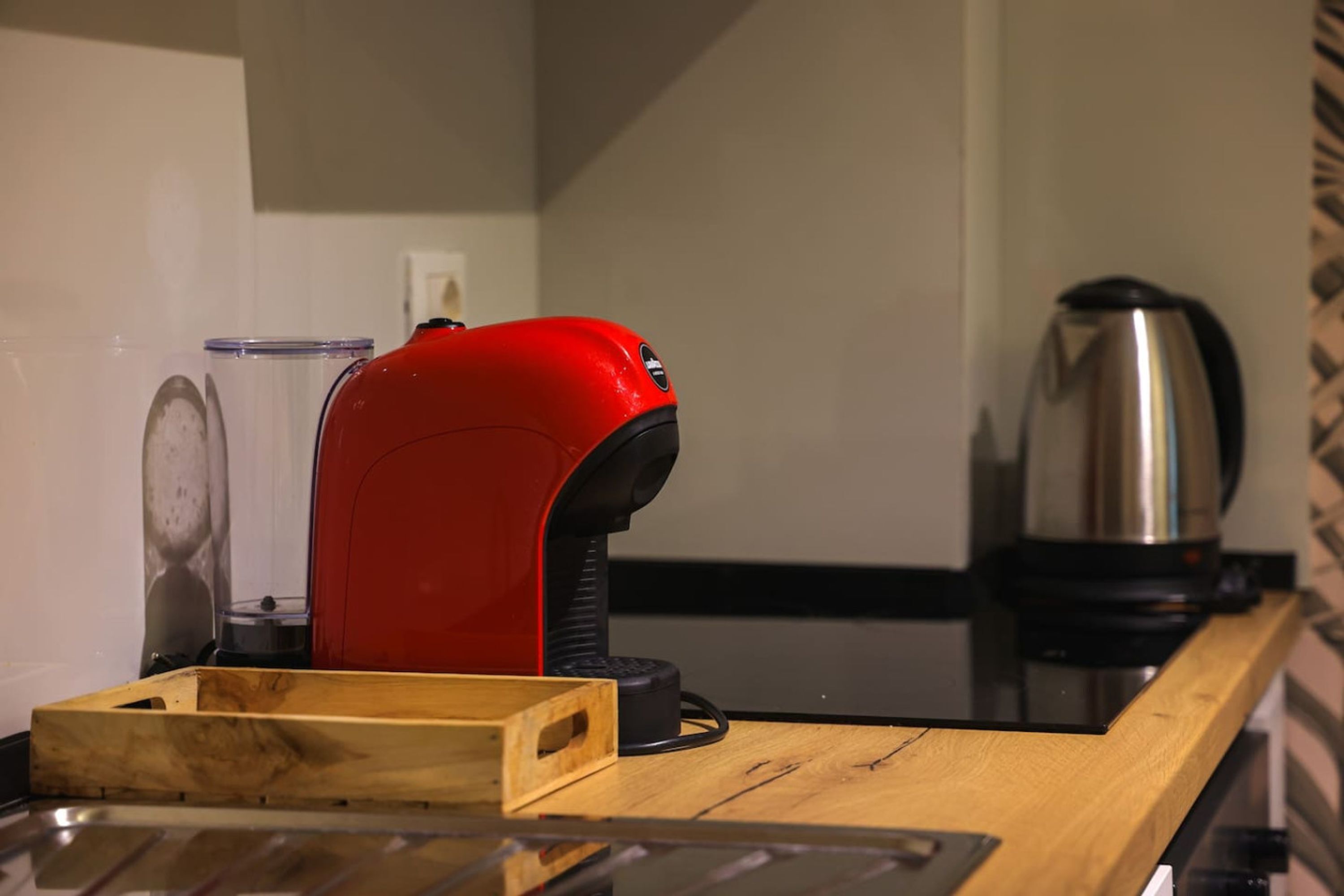 A kitchen counter with a red coffee machine, a stainless steel kettle, and a wooden tray.