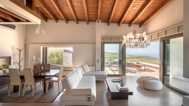 Ein geräumiges Wohnzimmer mit Holzbalkendecke, einem weißen Sofa, einem Essbereich und einer großen Glasschiebetür, die auf eine Terrasse im Freien führt.