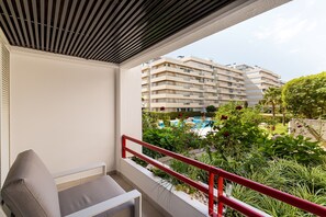 Ein Balkon mit roter Geländer, einer weißen Wand und Blick auf ein mehrgeschossiges Gebäude sowie ein Schwimmbecken.