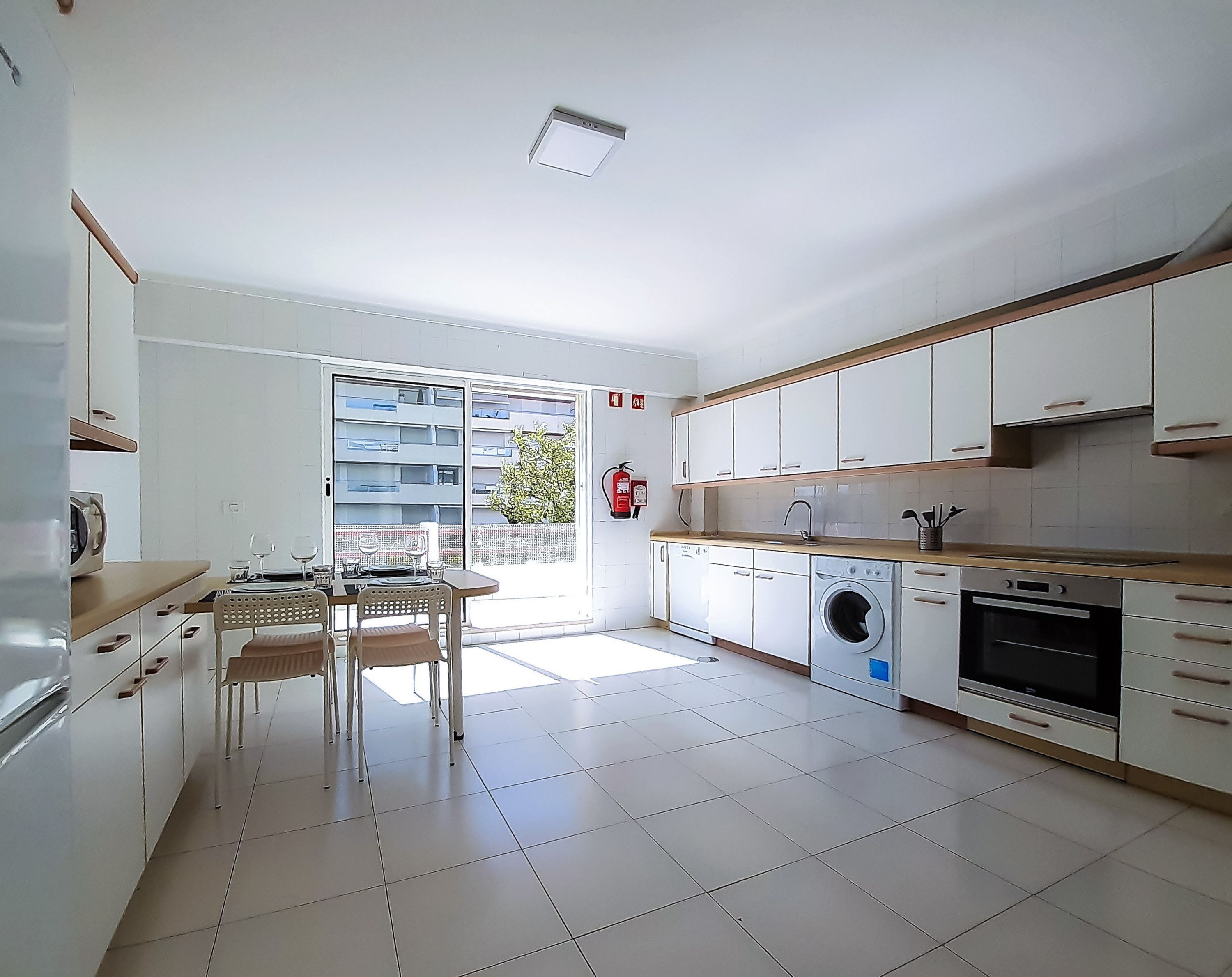 A modern kitchen with a built-in washing machine, a dining area with a table and chairs, and a sliding glass door leading to a balcony.