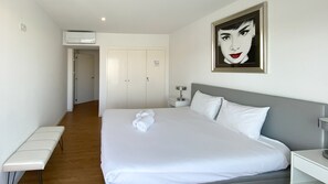 Hotelkamer met een bed, nachtkastje, een bankje, een wandairconditioner, een deur en een ingelijst kunstwerk.