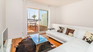 A modern living room with a white sofa, glass coffee table, and a view of the outside. - Onde a Marina é Casa | T2 HsRentals Vilamarina (Vilamoura)