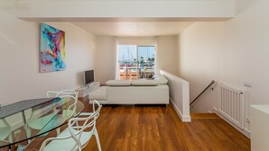 A modern living room with a glass table, white sofa, and a colorful abstract painting. - Onde a Marina é Casa | T2 HsRentals Vilamarina (Vilamoura)