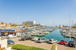 A marina with boats docked, a parking area with cars, and buildings in the background. - Onde a Marina é Casa | T2 HsRentals Vilamarina (Vilamoura)