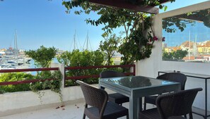A balcony with a glass table and wicker chairs, overlooking a marina with boats and a cityscape. - Vista Marina Exclusiva | T1 HsRentals Vilamarina (Vilamoura)