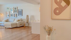 Ein modernes Wohnzimmer mit einer Couch, einem Couchtisch und einer Lampe. An den Wänden hängen gerahmte Kunstwerke, und auf einem Beistelltisch steht eine Vase mit getrockneten Blumen.