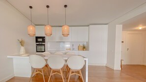 A modern kitchen with white cabinetry, a central island with four chairs, and three wicker pendant lights. - YES! Incrível Vista Piscina | T2 HsRentals Vilamar (Vilamoura)