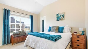 A bedroom with a large bed, a nightstand, a bench, and a window with curtains.