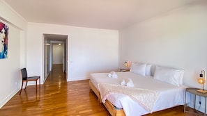 A hotel room with a large bed, bedside tables, a chair, and a hallway leading to another room. - Que sonho! Vista Marina | T2 HsRentals Vilamarina (Vilamoura)
