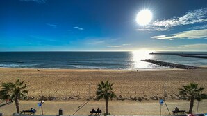 Ein Strand mit blauem Himmel, Sonne und ruhiger See. Palmen und ein Felssteg sind zu sehen.