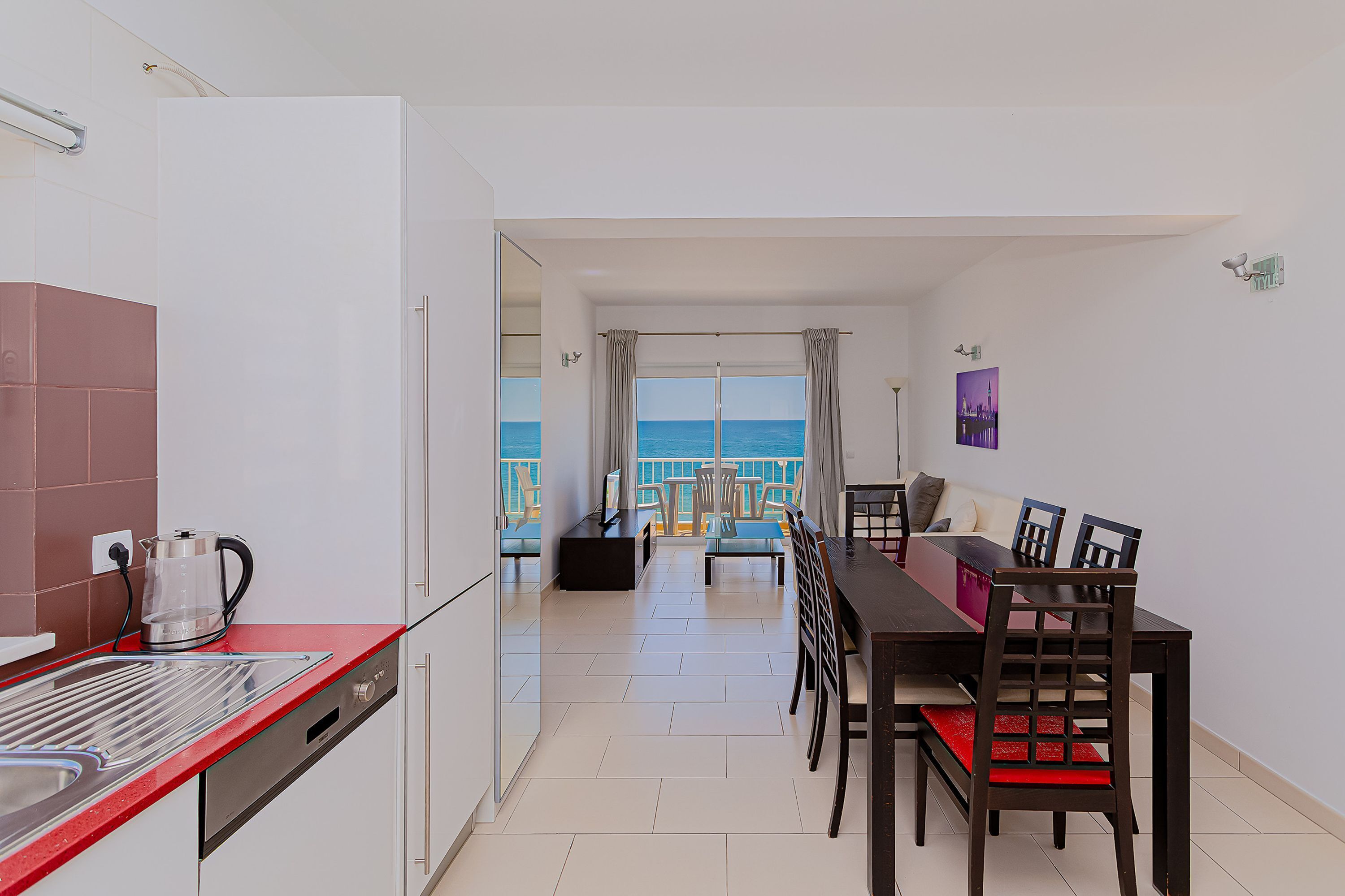 A modern kitchen with a red countertop, a coffee maker, and a dishwasher. There is a dining area with a table and chairs, and a view of the sea through large windows.