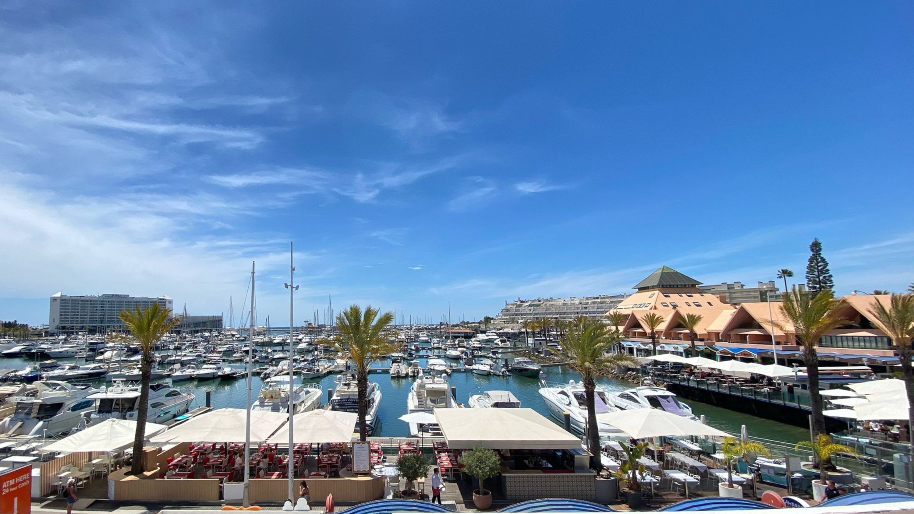 Un puerto deportivo con barcos, una zona de restaurantes y un hotel al fondo.