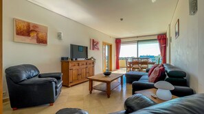 A living room with a sofa, a coffee table, a TV, and a view of a balcony with a table and chairs. - Aquamar Sunset | T2 HsRentals Marina Vilamoura (Vilamoura)